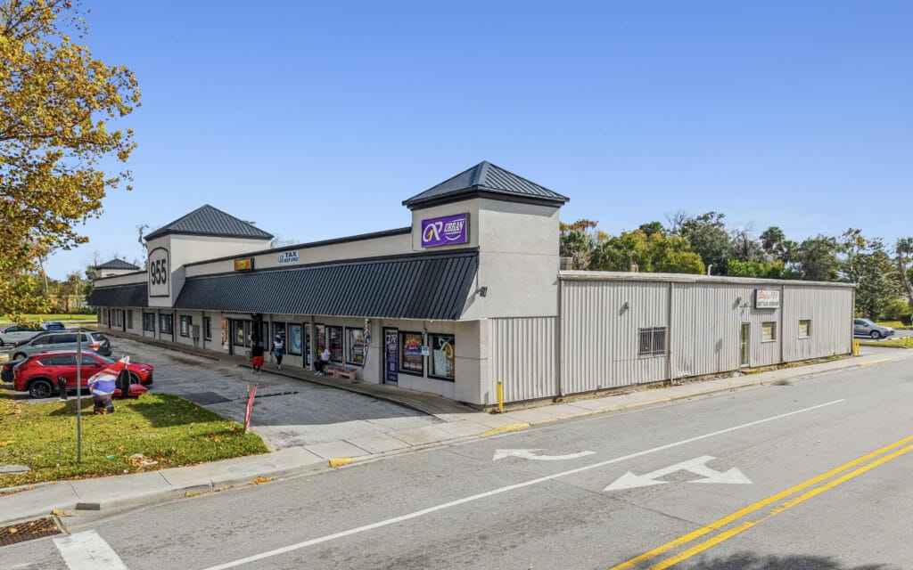 Exterior view of the Orange Shopping Center in Daytona Beach, featuring storefronts and a clear blue sky.